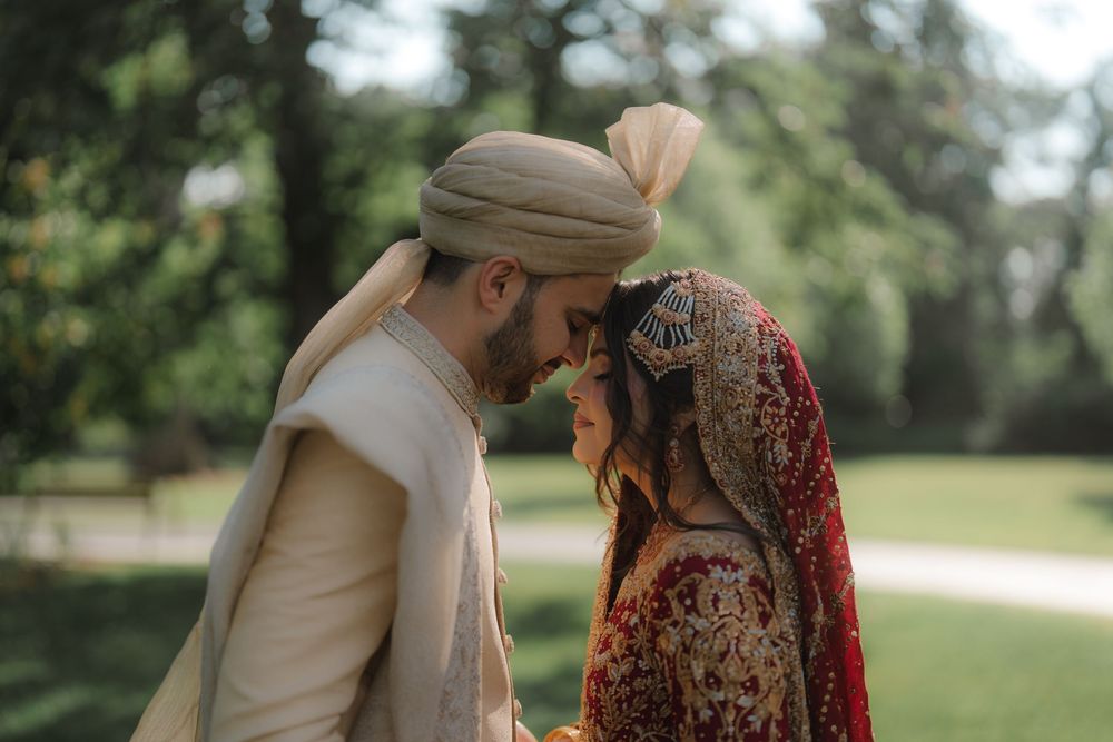 South Asian bride and groom in traditional wedding attire share an intimate moment outdoors amid lush green trees.