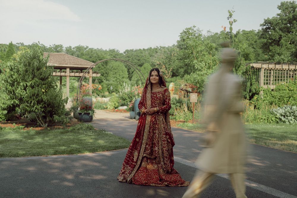 Bride in red bridal lehenga stands in lush garden as groom walks past in traditional South Asian wedding attire.