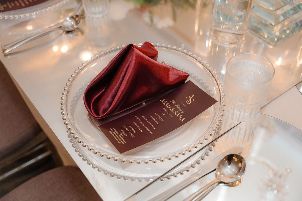 Elegant wedding place setting with burgundy napkin, beaded charger plate, menu card, and crystal glassware on white tablecloth.