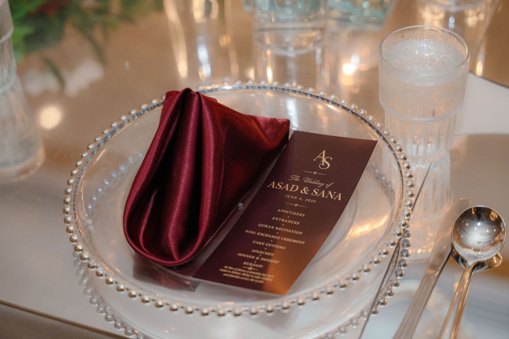 Elegant wedding place setting with beaded charger plate, burgundy satin napkin, and personalized Asad & Sana menu card.
