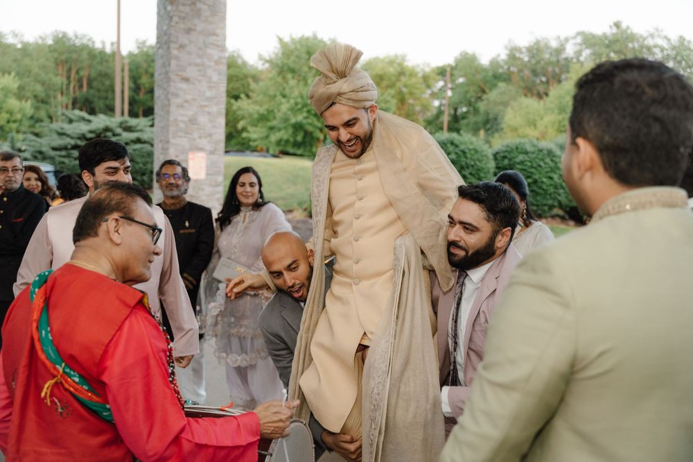 Groom in gold sherwani lifted by groomsmen during Indian wedding ceremony outdoors.