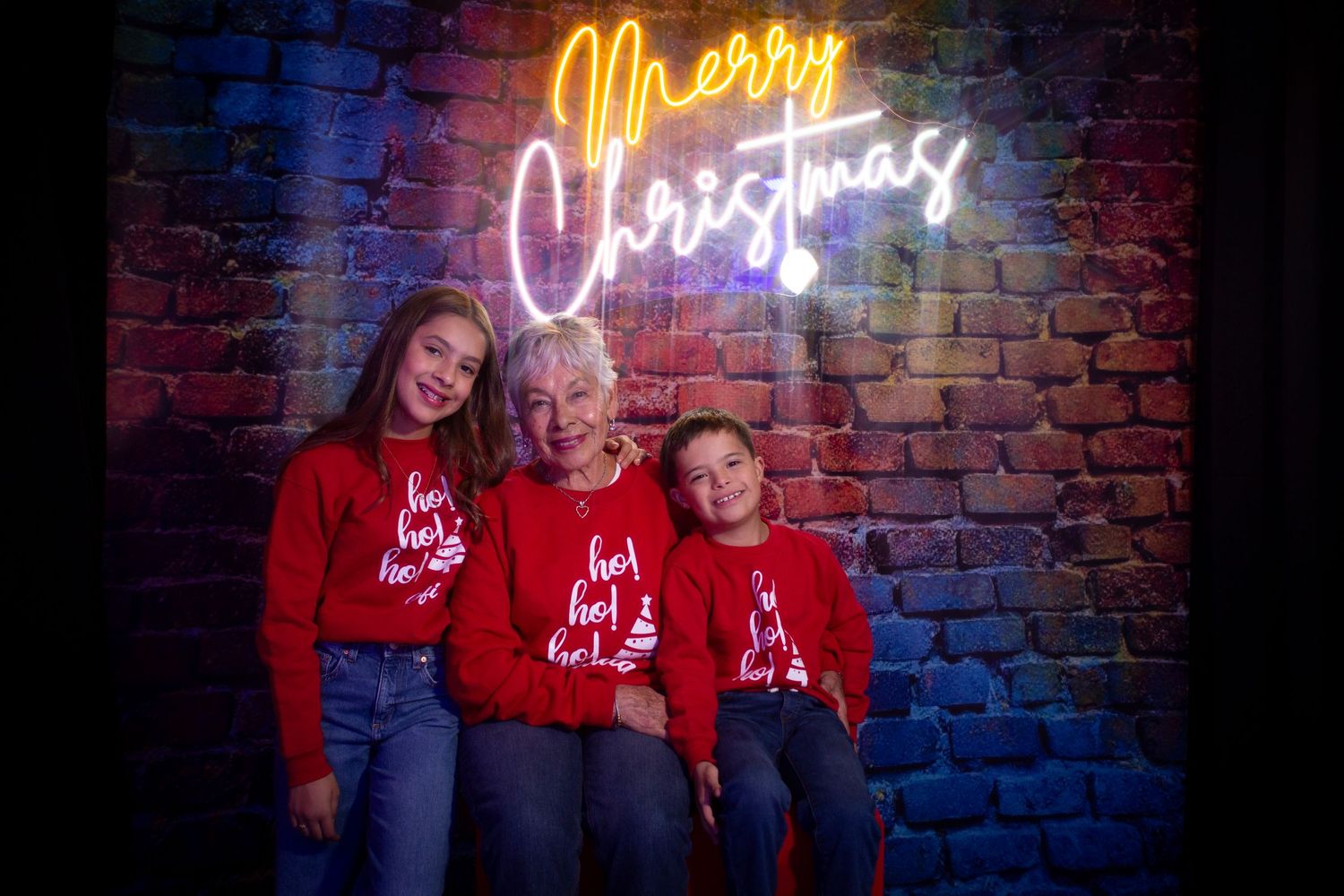A family poses for a holiday portrait in front of a brick wall with glowing neon 'Merry Christmas' sign.