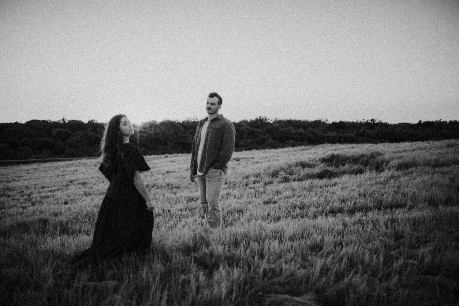 Black and white photo of a couple standing in a field at dusk, woman in dark dress, man in casual attire.