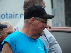 Older man in black cap and blue shirt standing outdoors at a public event.