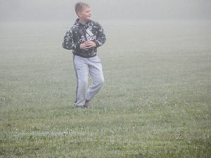 A young boy in a camo hoodie and gray sweatpants runs across a misty green field on a foggy day.