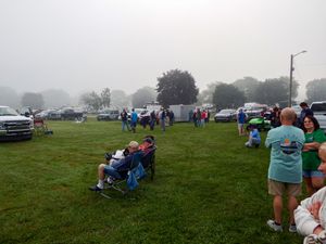 People gathering on a misty green lawn at an outdoor event with cars parked in the background on a foggy day.