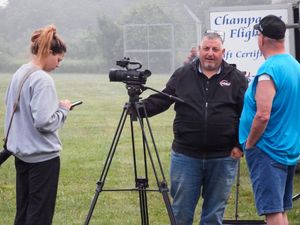 Outdoor video crew filming near a Champagne Flight sign, with a cameraman on tripod and woman taking notes.