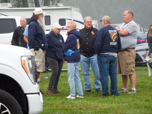 Group of adults in blue jackets gathered outdoors near vehicles at an event on a cloudy day.