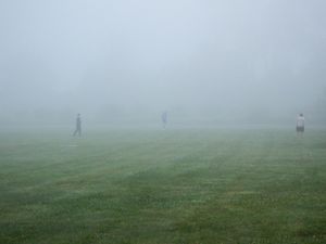 People walking on a foggy green field with low visibility and misty atmospheric conditions on a cloudy day.
