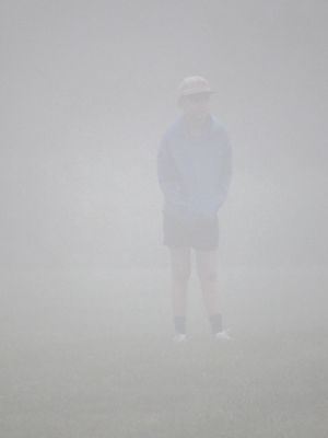 A solitary person stands shrouded in dense fog, wearing a hat and shorts, barely visible through the thick mist.
