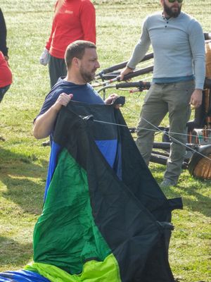 Man holding colorful paraglider canopy on green field with other participants preparing equipment outdoors.