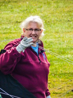 Smiling woman in burgundy fleece and gloves holding kite strings outdoors on green grass.