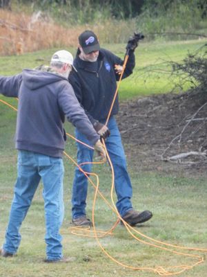 Two men untangle orange extension cords on a grassy field outdoors.