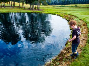 A young red-haired boy stands beside a calm reflective pond on a lush green golf course surrounded by tall trees.