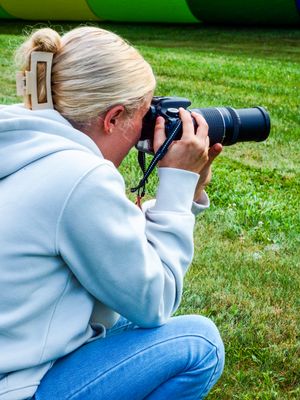 Blonde woman crouching on grass, photographing with a DSLR camera featuring a telephoto lens outdoors.