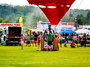 Hot air balloon at outdoor fair with food vendors selling meatball subs, snow cones, funnel cakes, and cotton candy.