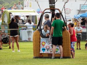People gather around a hot air balloon basket at an outdoor fair on a grassy field on a cloudy day.