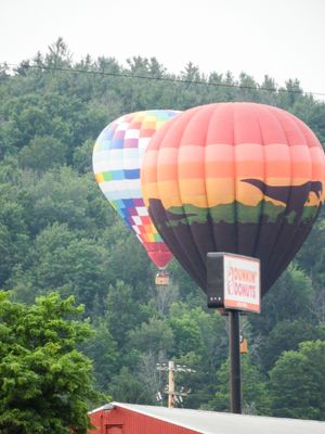 Two colorful hot air balloons float near a Dunkin Donuts sign with a lush green forested hillside in the background.