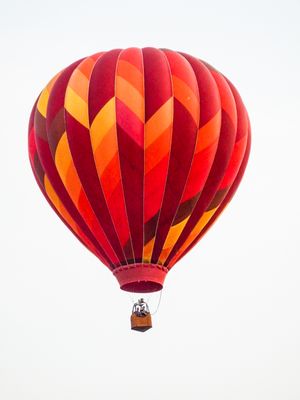 Vibrant red, orange, and yellow striped hot air balloon soaring against a clear white sky with passengers in the basket.
