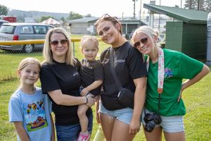 Group of five females posing outdoors at a summer fair, wearing casual black t-shirts, smiling together on green grass.