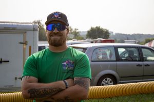 Bearded man in green t-shirt and sunglasses stands arms crossed near yellow hose outdoors at event.