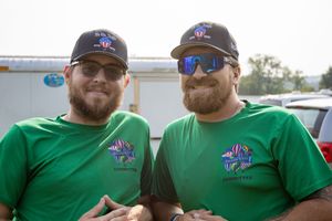 Two men wearing matching green shirts and black caps with sunglasses smile outdoors at an event.