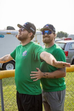 Two bearded men in matching green shirts and caps with sunglasses stand by a chain-link fence outdoors.
