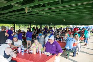 Large crowd gathered under a green pavilion shelter enjoying an outdoor community picnic event with colorful tablecloths.