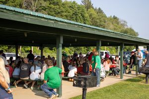 Large group of people gathered under a covered outdoor pavilion at a sunny park picnic event.