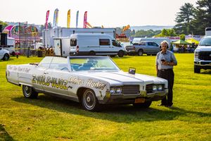 Vintage white convertible car with custom lettering parked on grass at an outdoor event with trailers in background.