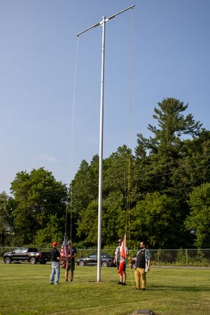 People raising American and state flags on a tall flagpole in a green field with trees in the background.