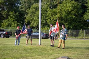 Four men holding American and Canadian flags on a grassy field near a flagpole during an outdoor ceremony.