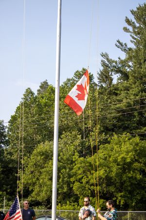 Canadian flag raised on a flagpole outdoors with people standing nearby and lush green trees in the background.