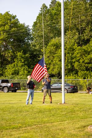 Two men raising an American flag on a flagpole in a green field with trees and parked cars in the background.