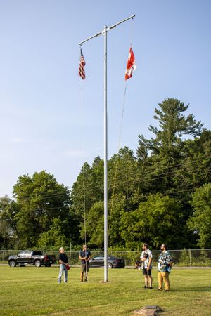 Four people stand near a tall flagpole flying American and red flags on a green lawn with trees in the background.