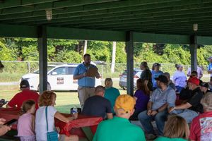 Man speaks at outdoor covered pavilion gathering with people seated at red picnic tables in summer.