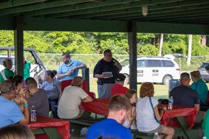 Two men present documents at an outdoor community meeting under a pavilion with attendees at picnic tables.