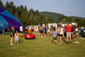 Crowd gathers on green field near colorful hot air balloon during outdoor festival with wooded hills in background.