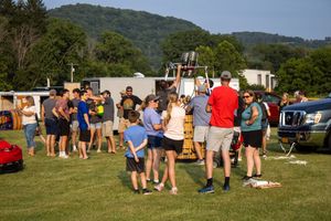 Group of people gathered outdoors on grass near vehicles and hills for a community event on a sunny day.