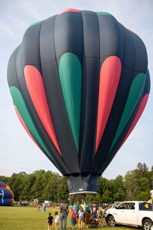 Dark hot air balloon with green and pink panels inflating at outdoor festival with crowd of spectators on green field.