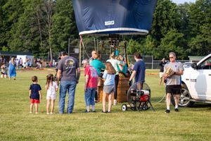 Families gather around a hot air balloon basket at an outdoor festival on a sunny day in a green park.