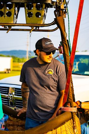 Man in baseball cap and sunglasses standing in a hot air balloon basket outdoors on a sunny day.