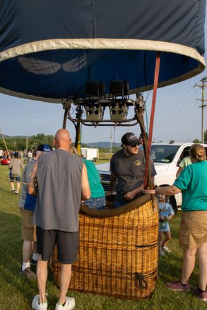 People gather around a wicker hot air balloon basket at an outdoor festival on a sunny day.