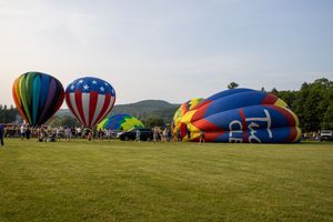 Hot air balloons in red, white, and blue patterns inflate on a green field at a balloon festival with mountains in the background.