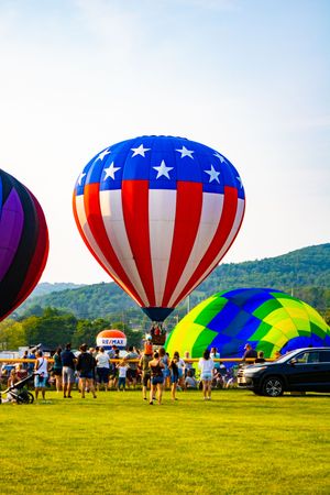 American flag hot air balloon soars at a colorful balloon festival, with crowds on green grass and mountains in the background.