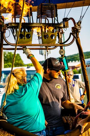 People operating a hot air balloon burner system in a wicker basket at an outdoor balloon festival event.