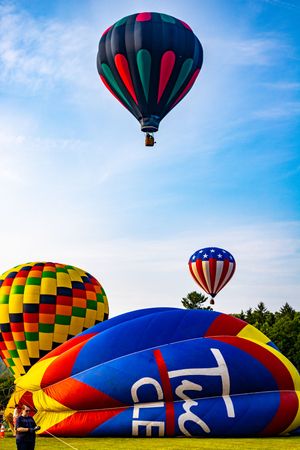 Colorful hot air balloons at a festival, with one patriotic and one dark balloon soaring above a green field under blue skies.