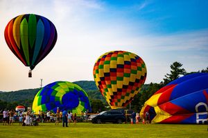 Colorful hot air balloons launching at a summer festival on a green field with mountains in the background.