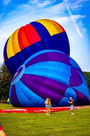 Two people stand beside colorful hot air balloons being inflated on a green field under a bright blue sky.