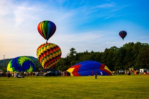 Colorful hot air balloons launching at a festival on a green field with trees and blue sky backdrop.